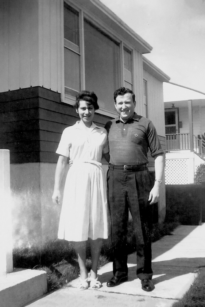 Photo en noir et blanc d'un homme et d'une femme souriant et se tenant debout dans l'allée à l'extérieur d'une maison. Il semble que ce soit l'été. La femme porte une robe à manches courtes.