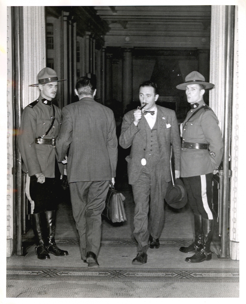 Photo en noir et blanc de quatre hommes dans une grande porte. Un homme, le deuxième à partir de la droite, sort de la pièce avec un attaché-case et a une pipe dans sa bouche. L'homme à sa gauche entre dans la pièce. Les deux hommes se tenant de part et d'autre portent un uniforme de la Gendarmerie royale du Canada et regardent en direction de la caméra.