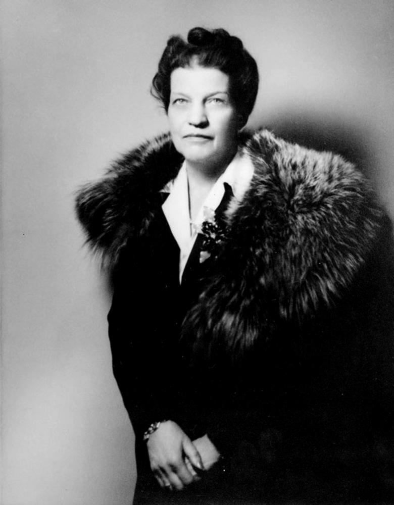 Photo en noir et blanc d’un portrait de femme à partir de la taille. Elle porte un manteau noir avec un col de fourrure. Ses cheveux sont courts et frisés et elle regarde directement la caméra.