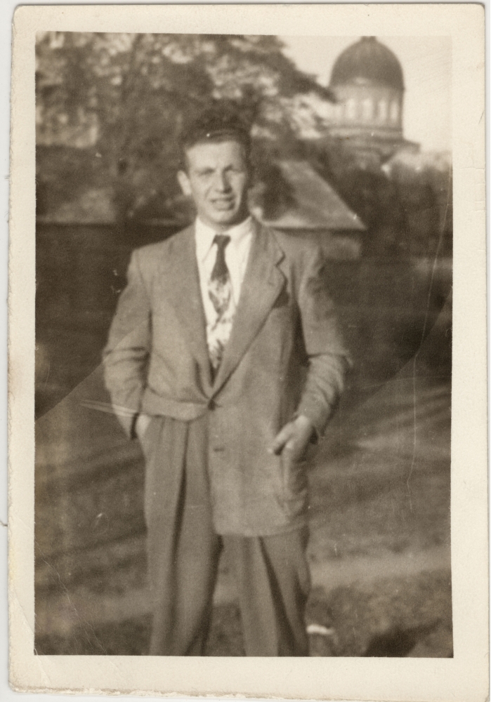 Black-and-white photograph of a man wearing a suit and tie, standing outdoors on a field. A building tower is visible in the background.