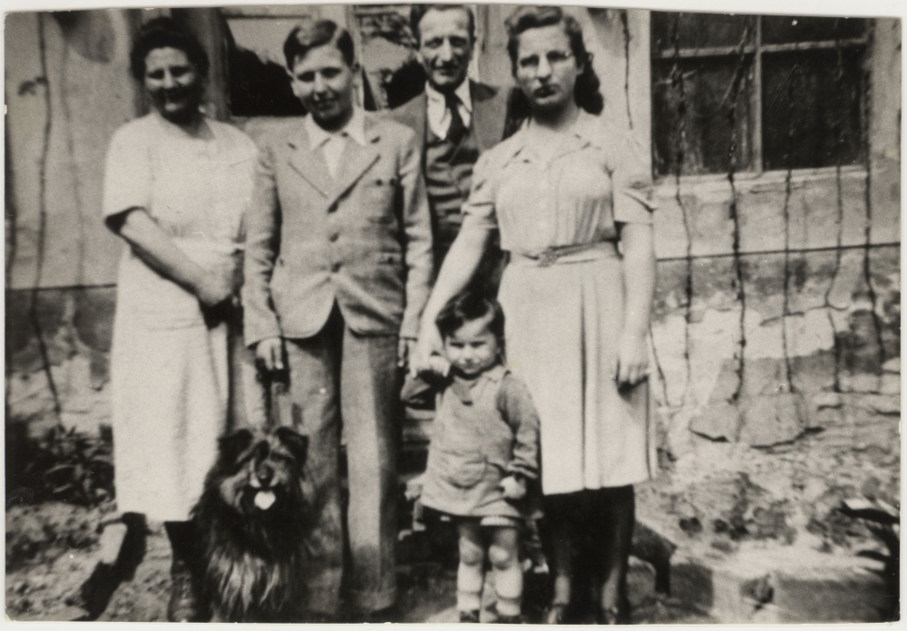 Photo en noir et blanc d'un homme et d'une femme debout à l'extérieur d'une maison avec deux adolescents, un jeune enfant et un chien.