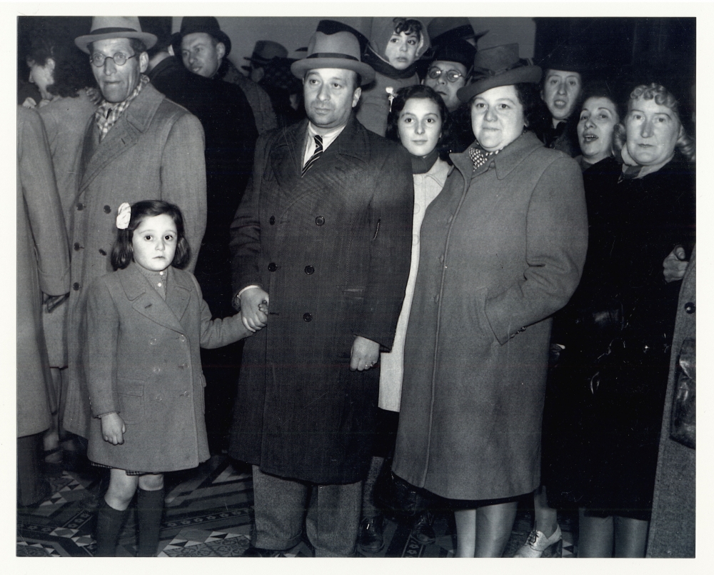 Photo en noir et blanc d'un groupe d'une douzaine de personnes rassemblés ensemble et regardant vers la caméra. Ils portent des manteaux et des chapeaux. L'homme dans la première rangée tient la main d'une jeune fille avec une boucle dans les cheveux.