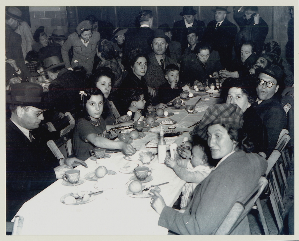 Black-and-white photograph of a large group of people sitting together at a long table, looking at the camera. The table is full of dishes and food. There are several more people in the room behind the table.