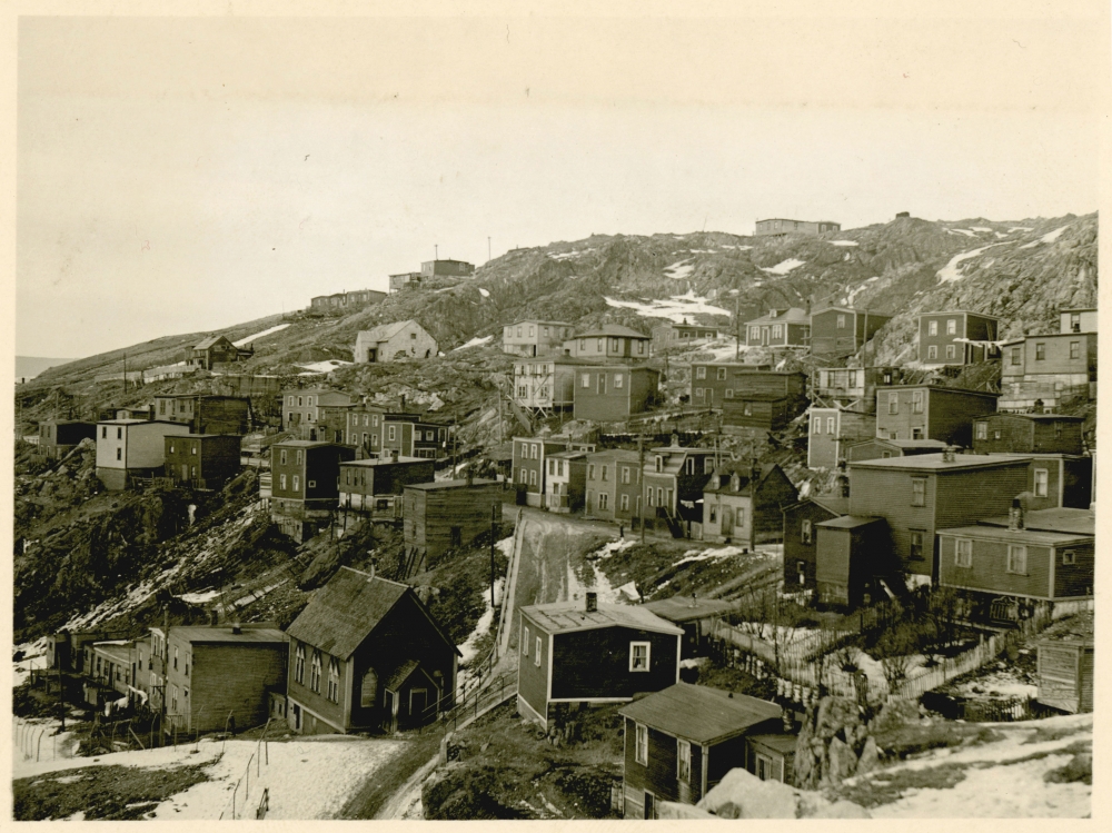 Black-and-white photograph of small residential buildings in a hillside town or village. There is snow on the ground and it appears to be winter.