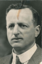 Black-and-white portrait photograph of a man, photo taken from his shoulders up. He is wearing a suit and tie.