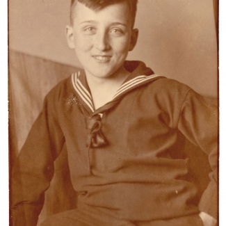 Sepia-toned photograph of an adolescent boy sitting and smiling at the camera. He wears a dark top with a sailor collar.