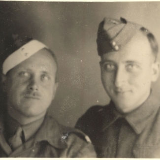 Black-and-white photograph of two men dressed in military uniform, smiling at the camera. The man on the left has a moustache.