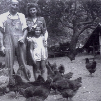 Black-and-white photograph of a man and woman with a young child with curly blond hair. They stand together outdoors in front of a truck and a barn in the background.