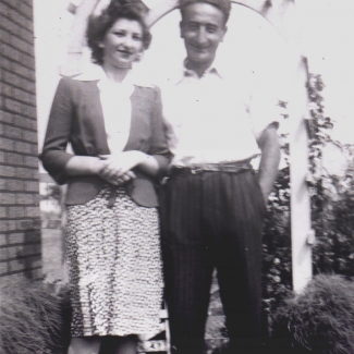 Black-and-white photograph of a man and woman standing together under an arbour outdoors, smiling at the camera. The man has his hands in his pockets.