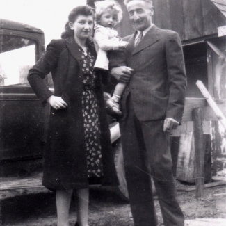 Black-and-white photograph of a man and woman with a young child with curly blond hair. They stand together outdoors in front of a truck and a barn in the background.