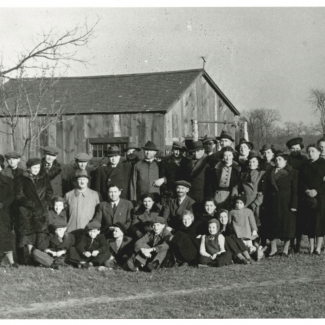 Photo en noir et blanc d'un grand groupe d'environ 35 hommes, femmes et enfants, habillés de manteaux d'hiver et de chapeaux, assemblés à l'extérieur sur une pelouse. Il y a une grange à l'arrière-plan derrière le groupe.