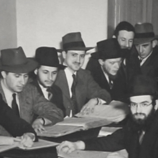 Photo en noir et blanc d'un groupe de dix hommes assis ensemble à une table couverte de livres et de papiers. Les hommes portent des costumes et des chapeaux.