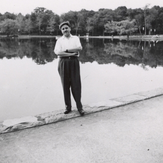 Photo en noir et blanc d'un homme se tenant debout les bras croisés devant un grand étang ou lac. Il porte une chemise à manches courtes, des pantalons noirs et un chapeau. Des arbres entourent le lac en arrière-plan.