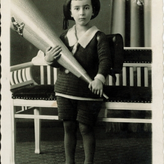 Portrait photographique pris en studio d'un jeune garçon en uniforme et chapeau d'écolier, regardant la caméra en adoptant une pose pensive. Il tient un gros objet ayant la forme d'un cornet. Il y a de l'écriture en allemand au bas de l'image, et les bords de la photo sont usés.