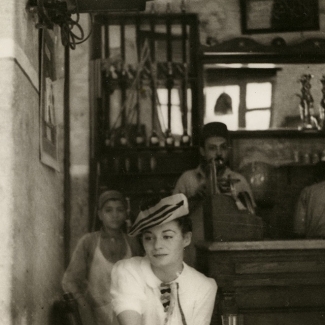 Black-and-white photograph of a woman sitting in a café, wearing a white blouse and beret. The woman is looking towards the side. A young boy and two men stand behind the bar in the background.