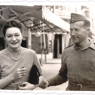 Black-and-white photograph of a woman and man outdoors. The woman holds a clutch purse under her arm and faces the camera, smiling. The man looks at the woman and holds her elbow.