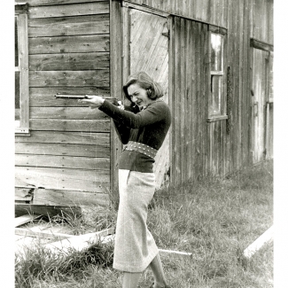 Photo en noir et blanc d'un femme debout devant une grange. Elle tient un fusil et le pointe à gauche de la caméra.