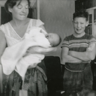 Photo en  noir et blanc d'un jeune garçon debout les bras croisés, souriant aux côtés d'une femme qui tient un bébé enveloppé dans une couverture.