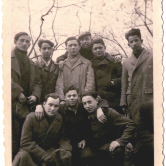 Black-and-white photograph of a group of 9 young men posing together arm-in-arm outdoors. They wear coats and there are bare trees above them in the background.