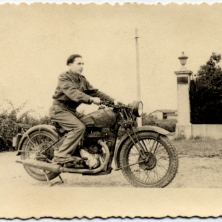 Black-and-white photograph of a young man posing on the back of a motorcycle, looking forwards towards the right of the camera. There is a hedge behind him and a building in the distant background.