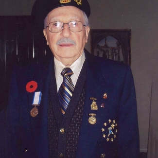 Colour photograph of an elderly man dressed in his military uniform, smiling at the camera. His jacket and beret are adorned with medals and pins, as well as a Remembrance Day poppy.