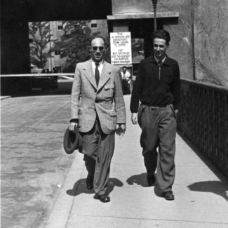 Black-and-white photograph of two men smiling and walking towards the camera on the sidewalk of a bridge. The man on the left we