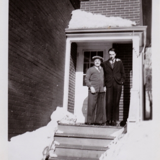 Black-and-white photograph of two men standing on the front porch of a brick house with snow in the foreground. Both men wear su