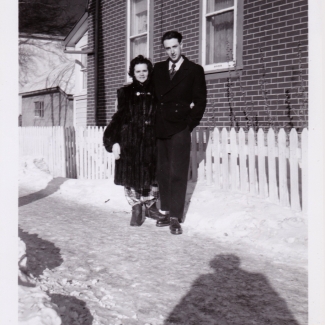 Black-and-white photograph of a woman and man standing together outdoors on a snowy driveway in front of a white fence. The woma