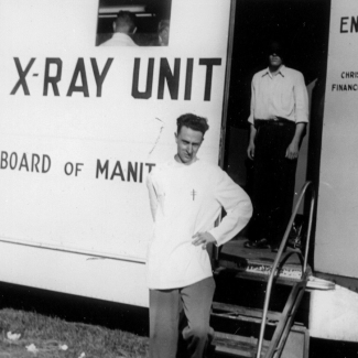 Black-and-white photograph of a man, standing with a hand on his left hip, in front of a large white truck that reads “X-RAY UNIT” on its side. A man stands behind him inside the entrance of the truck with his face covered by shadow.