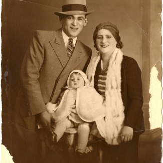 Photo de couleur sépia d'un homme et d'une femme prenant la pose avec un bébé pour un portrait. L'homme porte un chapeau ainsi que costume et cravate, et la femme porte aussi un chapeau. Le bébé porte une cape blanche. Les bords de la photo sont un peu usés et déchirés.