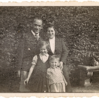 Photo en noir et blanc d'une femme et d'un homme debout ensemble avec deux jeunes enfants à l'extérieur, devant de grandes haies qui prennent tout l'espace en arrière-plan.