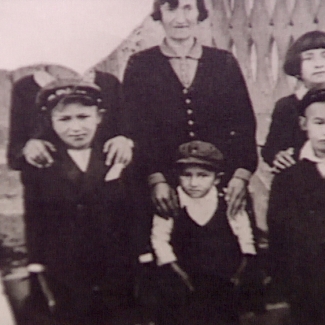 Black-and-white photograph of a woman with five children standing together, the three in the back row putting their hands on their family members’ shoulders in front. The photograph is torn in the top-left corner so that the face of the person standing in the top left is not visible.