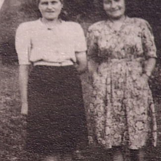 Photo en noir et blanc de deux jeunes femmes debout à l'extérieur et souriantes. Une des femmes porte une robe et l'autre une blouse et une jupe.