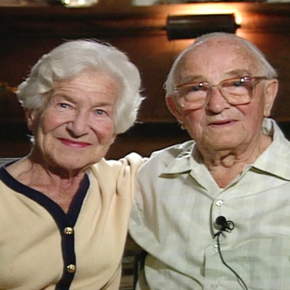 Photo en couleur d'un couple de personnes âgées assis ensemble bras-dessus bras-dessous et souriant à la caméra. La femme sur la gauche porte un cardigan et l'homme porte des lunettes et une chemise à col.