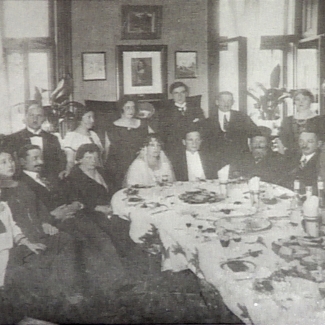 Black-and-white photograph of a group of about 20 people sitting and standing around a large dining table set with dishes. The group is formally dressed, and a couple sitting at the table appear to be celebrating their wedding.