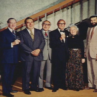 Colour photograph of a group of eight people standing together indoors in a line, smiling at the camera. The seven men wear suits, and the woman in the group wears a long dress.