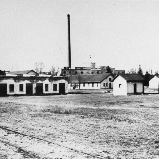 Black-and-white photograph of a field with one-storey barracks and a factory in the far background.