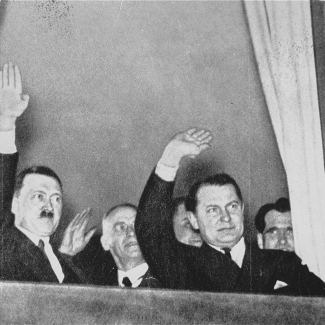 Photo en noir et blanc de deux hommes saluant à partir d'une fenêtre. La photo est prise d'en bas de l'extérieur. L'homme à gauche porte une moustache, et il y a des hommes derrière eux.