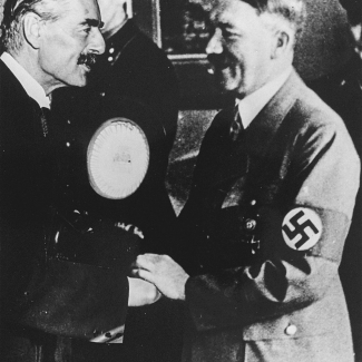 Black-and-white photograph of two men smiling and shaking hands. Both men have moustaches, and one man wears a lighter suit with a swastika badge on his arm.