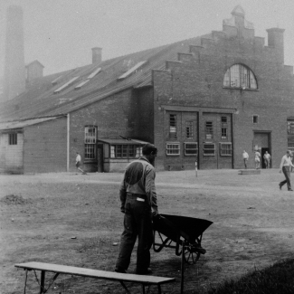 Photo en noir et blanc d'un homme, se tenant dos à la caméra, pousse une brouette dans une cours. Il y a un banc en avant-plan et un bâtiment en arrière-plan. L'homme porte un uniforme avec un grand cercle dans le dos.