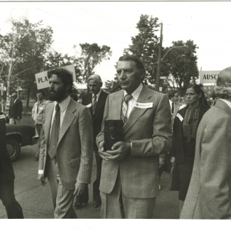 Photo en noir et blanc de quatre hommes menant une marche à l'extérieur. L'homme au centre transporte une boîte rectangulaire. Un groupe de personnes les suit à l'arrière.