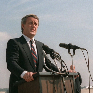 Colour photograph of a man in a suit, speaking outdoors at a podium hooked up with two microphones.