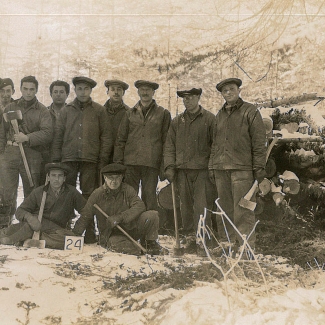 Black-and-white photograph of a group of eleven men posing together for a photograph in the woods. Some of the men are holding axes. They are grouped together in front of a pile of wood, wearing matching outdoor uniforms.