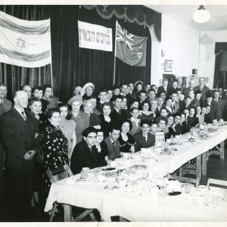 Black-and-white photograph of a large group of approximately 70 people smiling for a group photograph in a room. The group poses behind a large table with food and dishes on it, and there are two flags hung on curtains in the background.