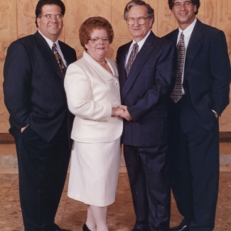 Colour studio photograph of a group of four adults standing together and smiling at the camera. The couple in the middle is elderly, and the woman wears a white jacket and skirt. The three men wear suits.