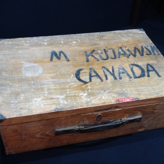 Colour photograph of a rectangular wooden suitcase with the words ‘M KUJAWSK, CANADA’ painted on the top of the case.