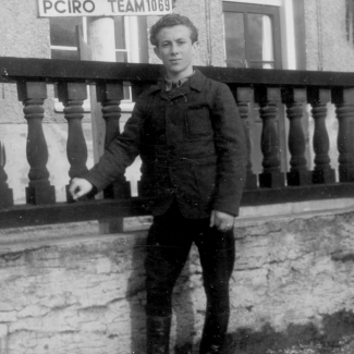 Black-and-white photograph of a young man standing outdoors on the grass and leaning against a raised  wooden railing behind him. A sign in the background reads ‘INTERNATIONAL CHILDRENS CENTER’.