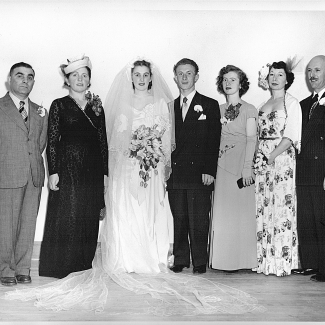 Photo en noir et blanc d'un groupe de sept personnes, se tenant en ligne et souriant à la caméra. Il semble que le couple au centre célèbre leur mariage puisque la femme est vêtue d'une longue robe blanche, d'un voile et tient un bouquet de fleurs. Les hommes du groupe portent des complets et les trois autres femmes des robes.