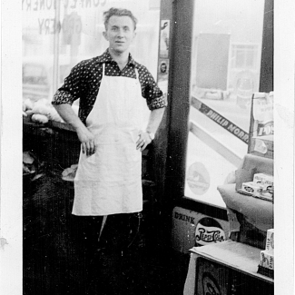 Black-and-white photograph of man standing inside a grocery store. He has his hands on his hips and wears a white apron, standing near the entrance door of the store. The window behind him is printed backwards with the words ‘LYONS CONFECTIONARY STORE’.