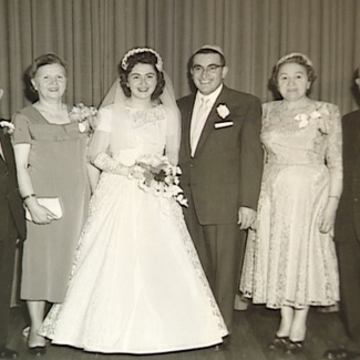 Photo en noir et blanc d'un groupe de six personnes debout en ligne et souriant à la caméra. Il semble que ce soit le mariage du couple du centre dont la femme porte une longue robe blanche, un voile et tient un bouquet de fleurs. Les trois hommes portent des complets et les deux autres femmes des robes.